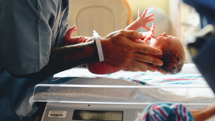A neonatal nurse carefully holds a premature infant in the NICU, representing the critical care needed for babies born disproportionately in disadvantaged communities.
